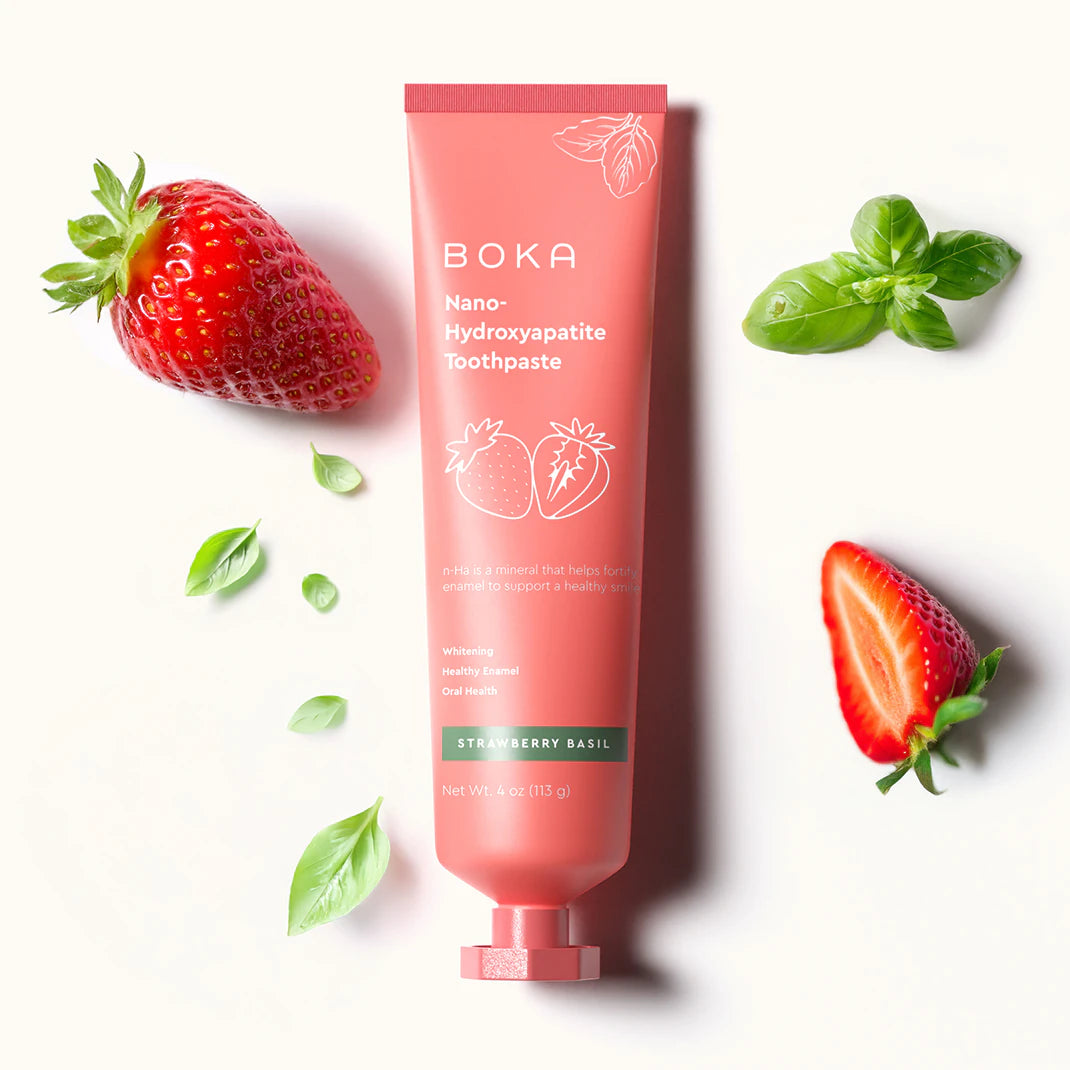 A pink tube of Boka toothpaste with strawberries and basil leaves on a white background.