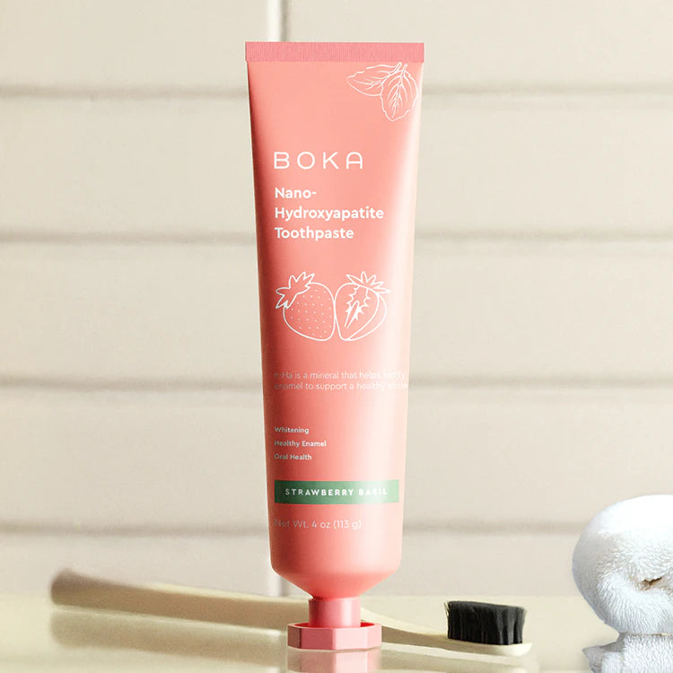 Pink tube of Boka Nano-Hydroxyapatite Strawberry Basil toothpaste standing upright by a toothbrush.