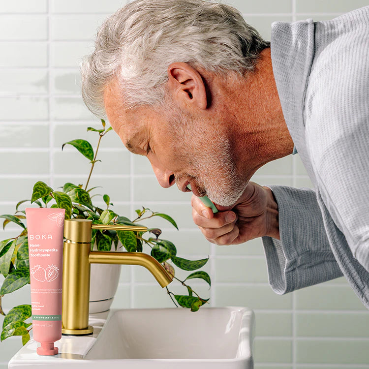 Older man in a robe brushes teeth at sink with pink Boka toothpaste, plants in background.