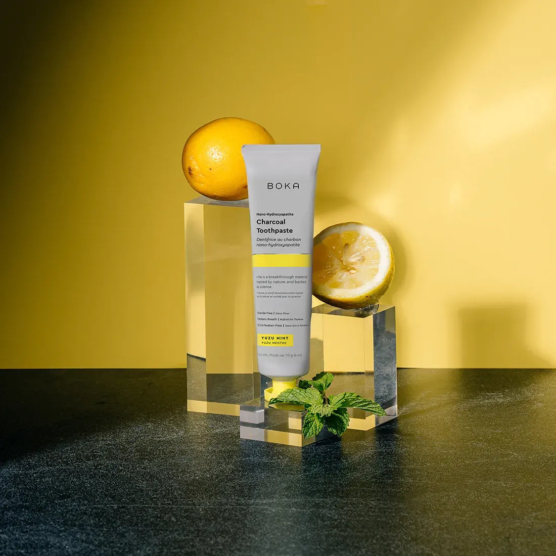 Boka charcoal toothpaste tube with lemon and orange on a reflective surface against a yellow background