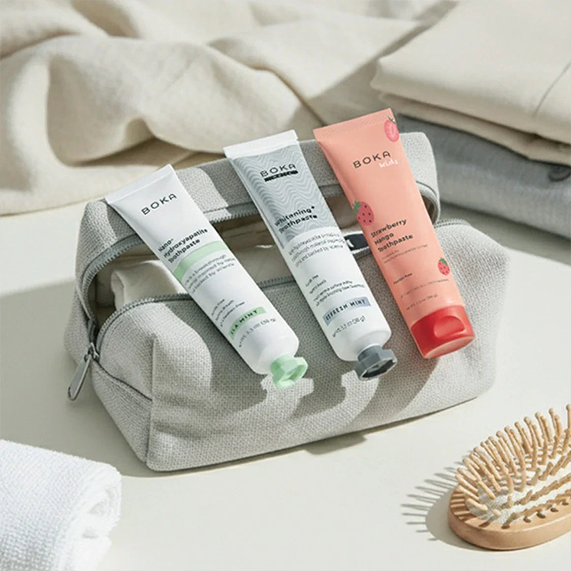 Three tubes of Boka toothpaste in a gray pouch with a wooden brush on a light background