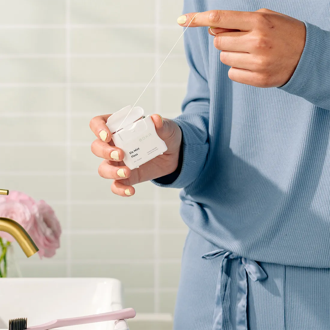 Person holding a Boka Ela Mint Floss, wearing a blue robe in a bathroom setting.