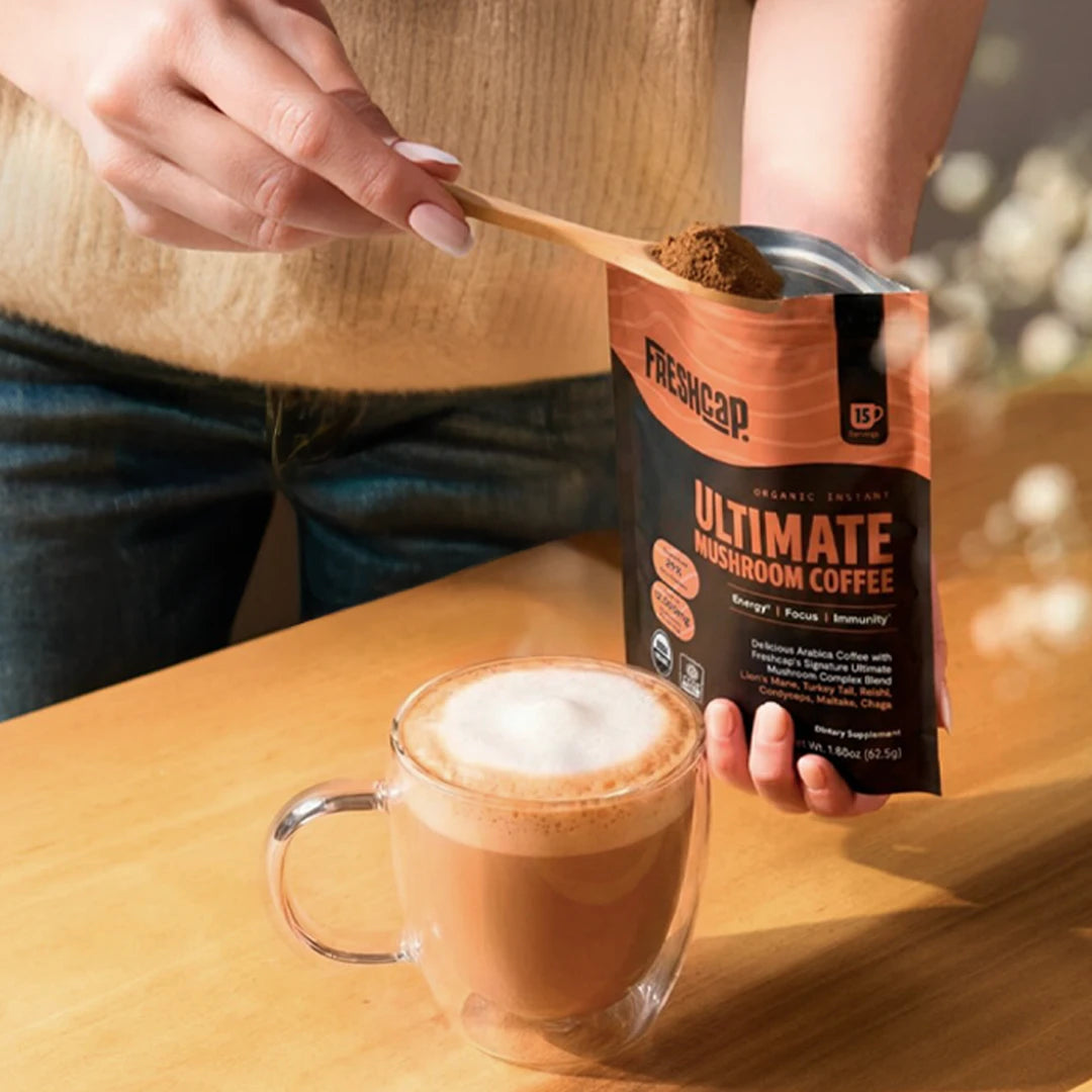 Person holding a can of Freshcap Ultimate Mushroom Coffee with a cup of coffee on a wooden table.