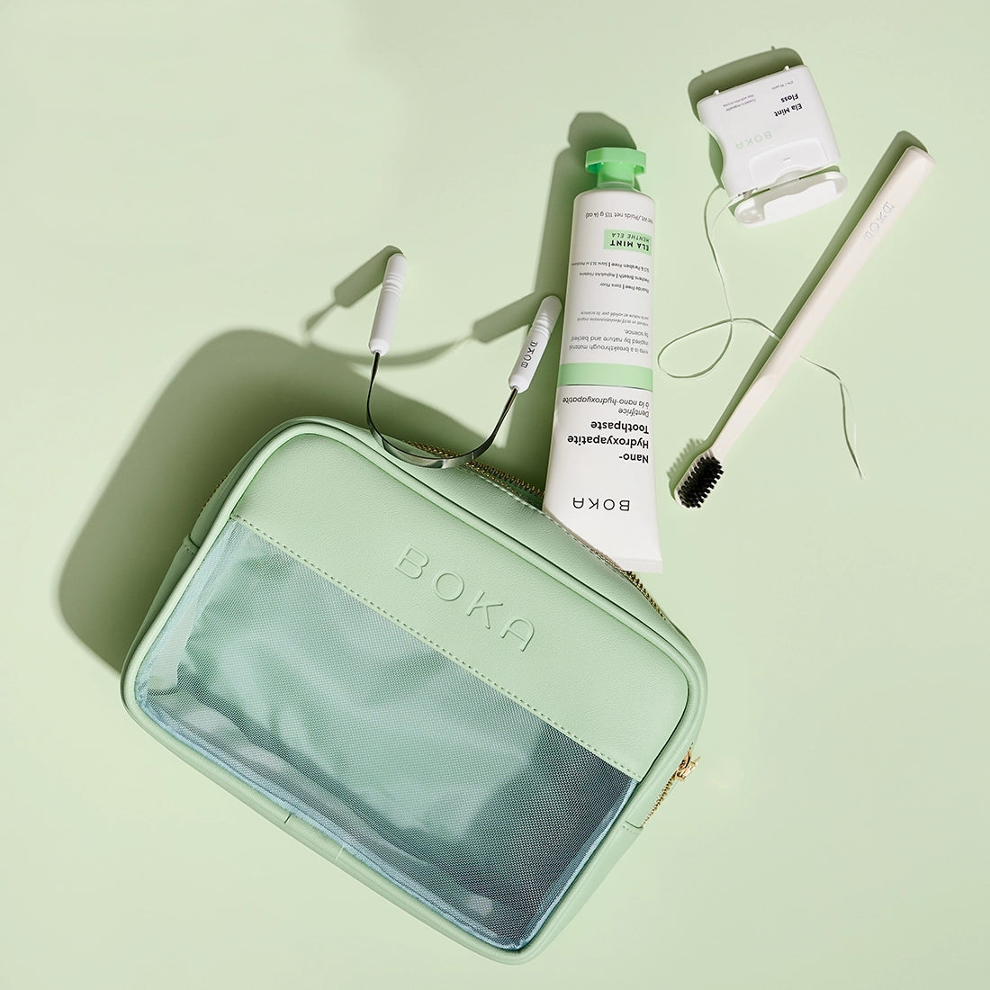 Mint green travel case with Boka branding, including a tube of cream, hairbrush, and other items on a light green background.