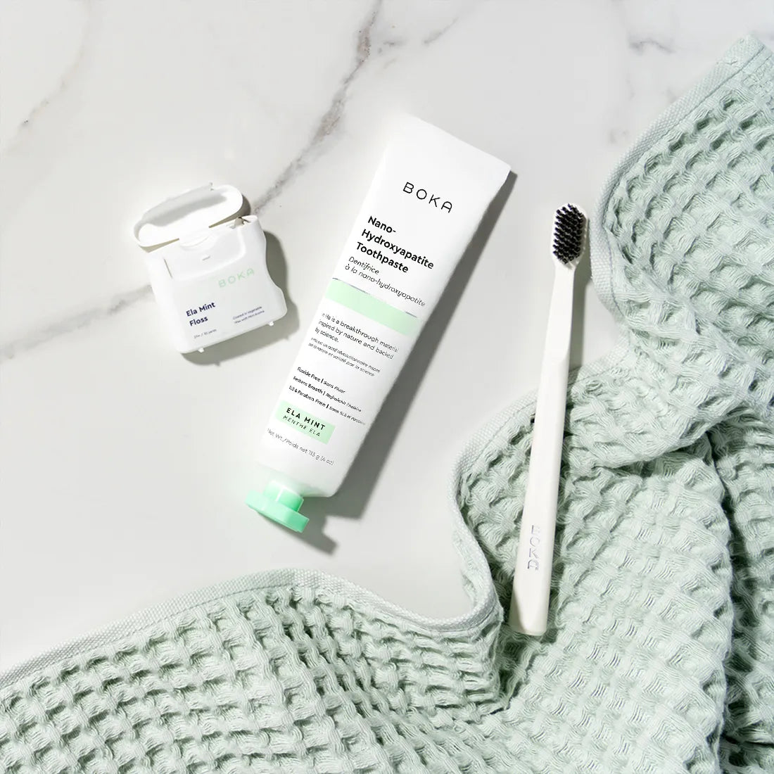 Boka toothpaste tube, toothbrush, and small container on a marble surface with a textured green towel.