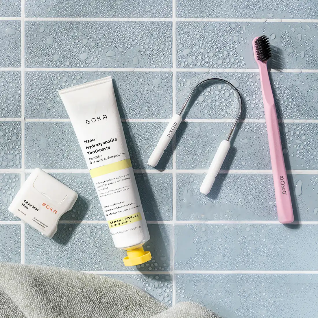 Boka toothpaste tube, brush, and cleaning tool on a tiled bathroom floor.