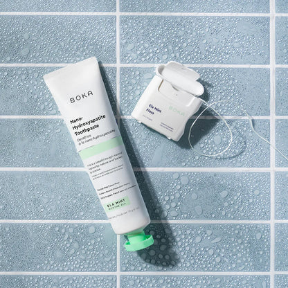 Boka toothpaste tube and dental floss on a tiled surface with water droplets.