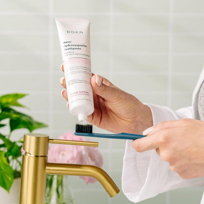 A person squeezes Boka activated charcoal toothpaste onto a Classic Toothbrush 2-Pack by a gold sink.