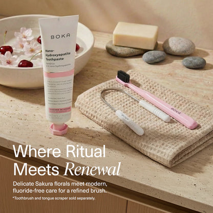 Boka toothpaste tube with a pink toothbrush and tongue scraper on a wooden surface with cherry blossoms.